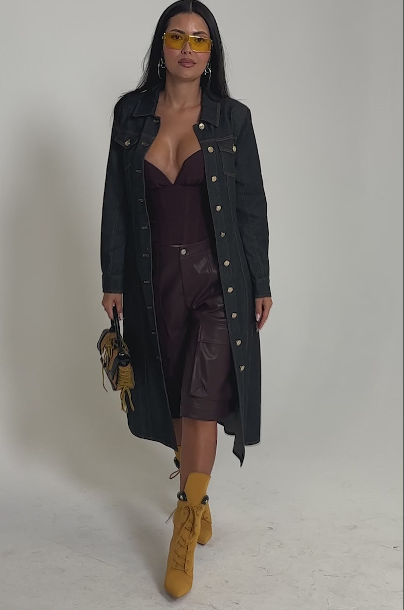 Video of woman modeling the ON YOUR RADAR DENIM TRENCH—a long-sleeve, belted, button-down dark-wash denim coat with gold stitching—paired with yellow heeled lace-up boots, yellow tinted sunglasses, and a small handbag.