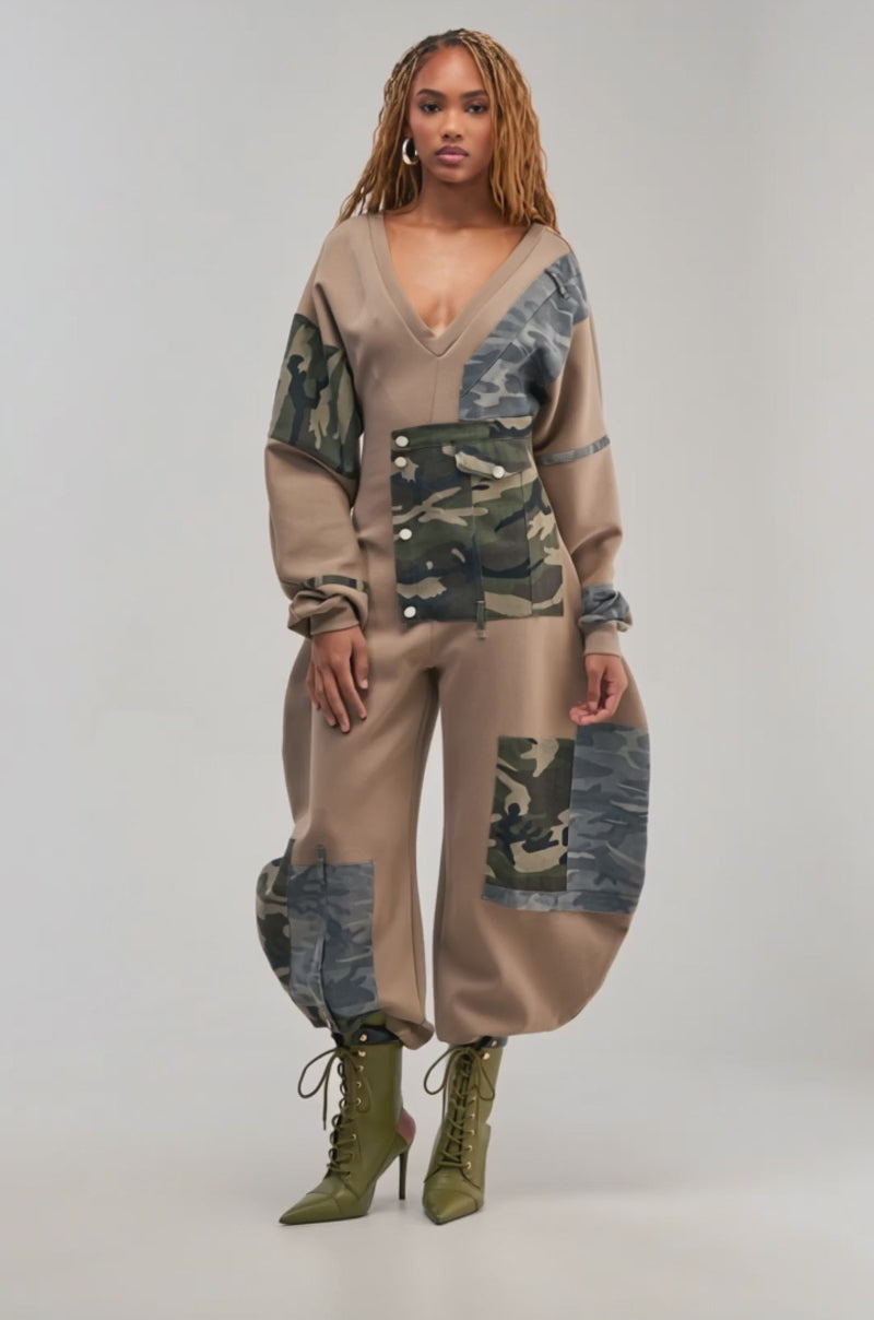 Video of a woman modeling the COMFY COZY PATCHWORK JUMPSUIT IN CAMO—featuring tan and camo details, loose legs, and long puffed sleeves.