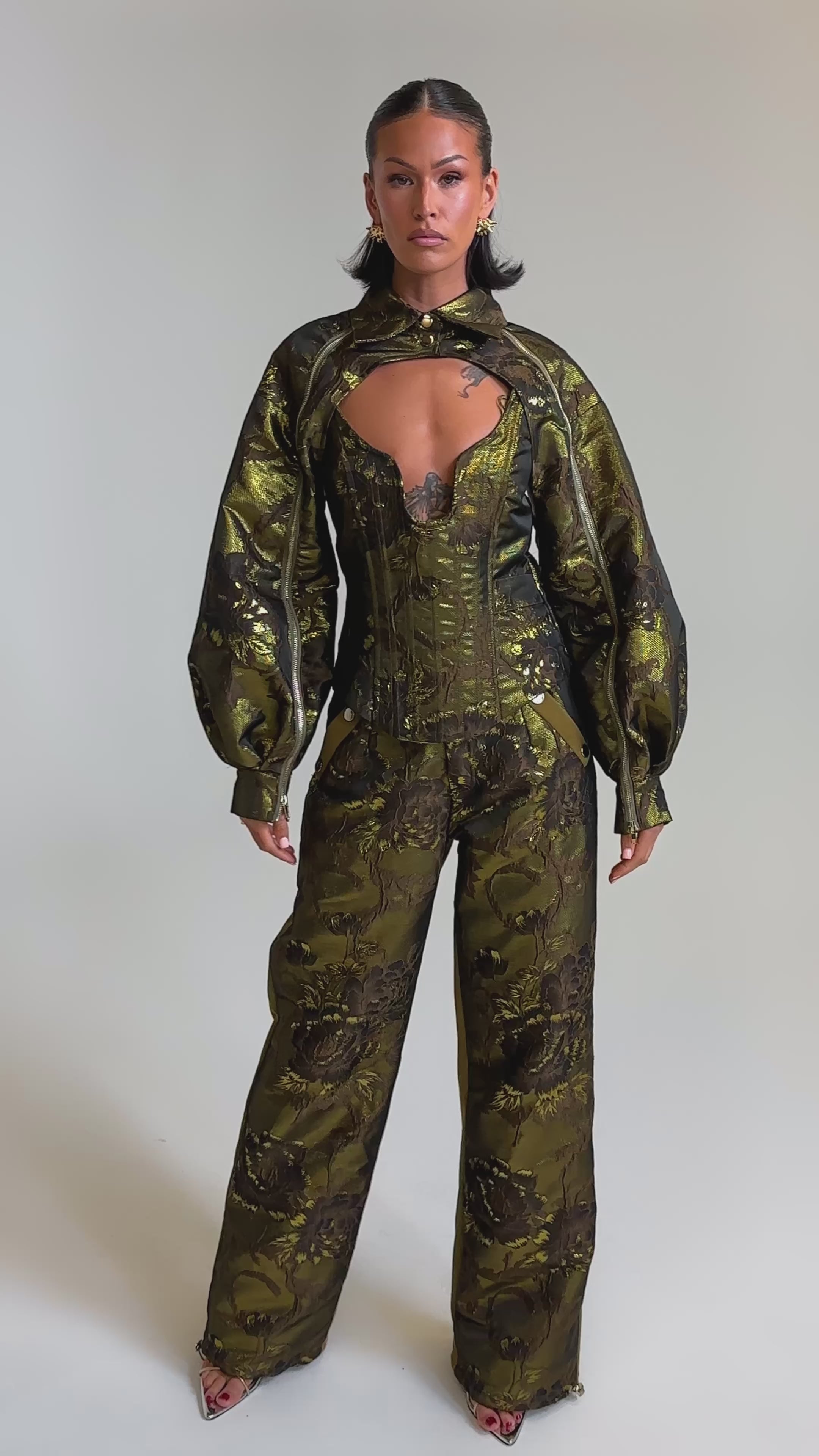 A video of a woman in the SHIGO BROCADE CORSET, styled with a fitted, patterned green and brown jumpsuit featuring voluminous sleeves and open-toe heels against a plain light background.