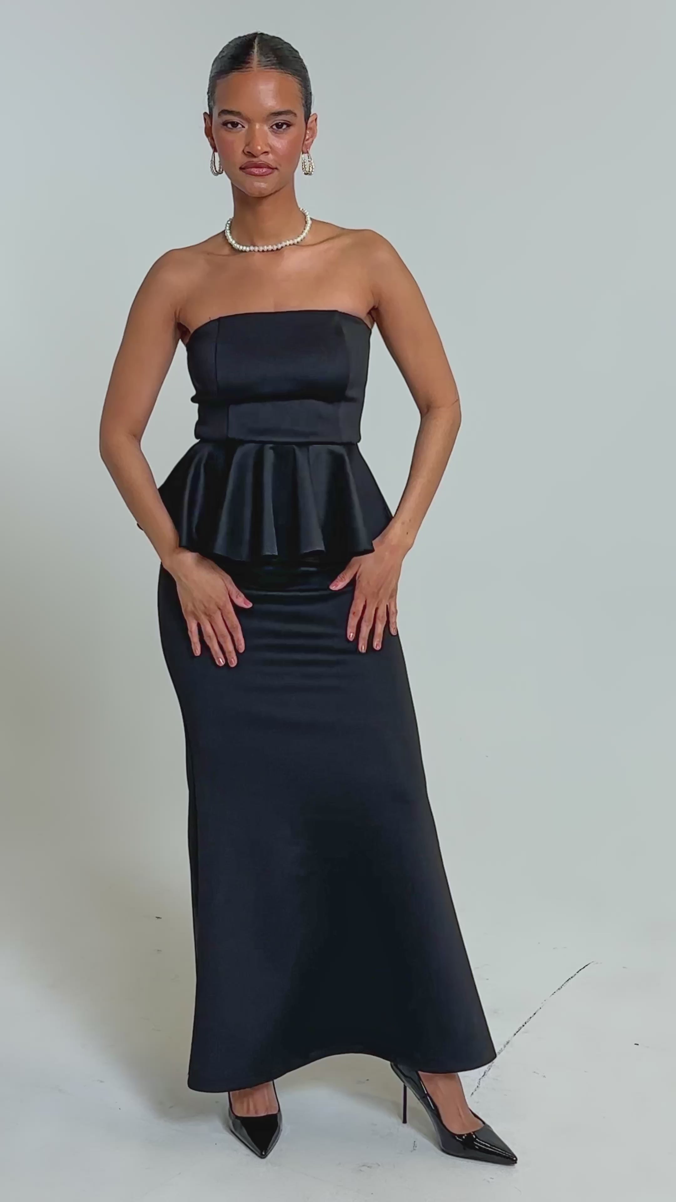 Video of woman modeling the BUILDING AN EMPIRE STRAPLESS MAXI DRESS, featuring a peplum waist and long fitted skirt. She completes this black eveningwear look with black heels, earrings, and a necklace against a plain light background.