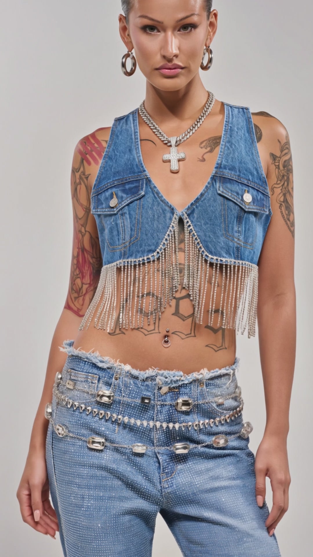 Video of woman modeling the RIDE EM CROPPED DENIM VEST as a crop top with matching denim pants, accessorized with hoop earrings, layered necklaces, and visible tattoos, posing against a plain background.