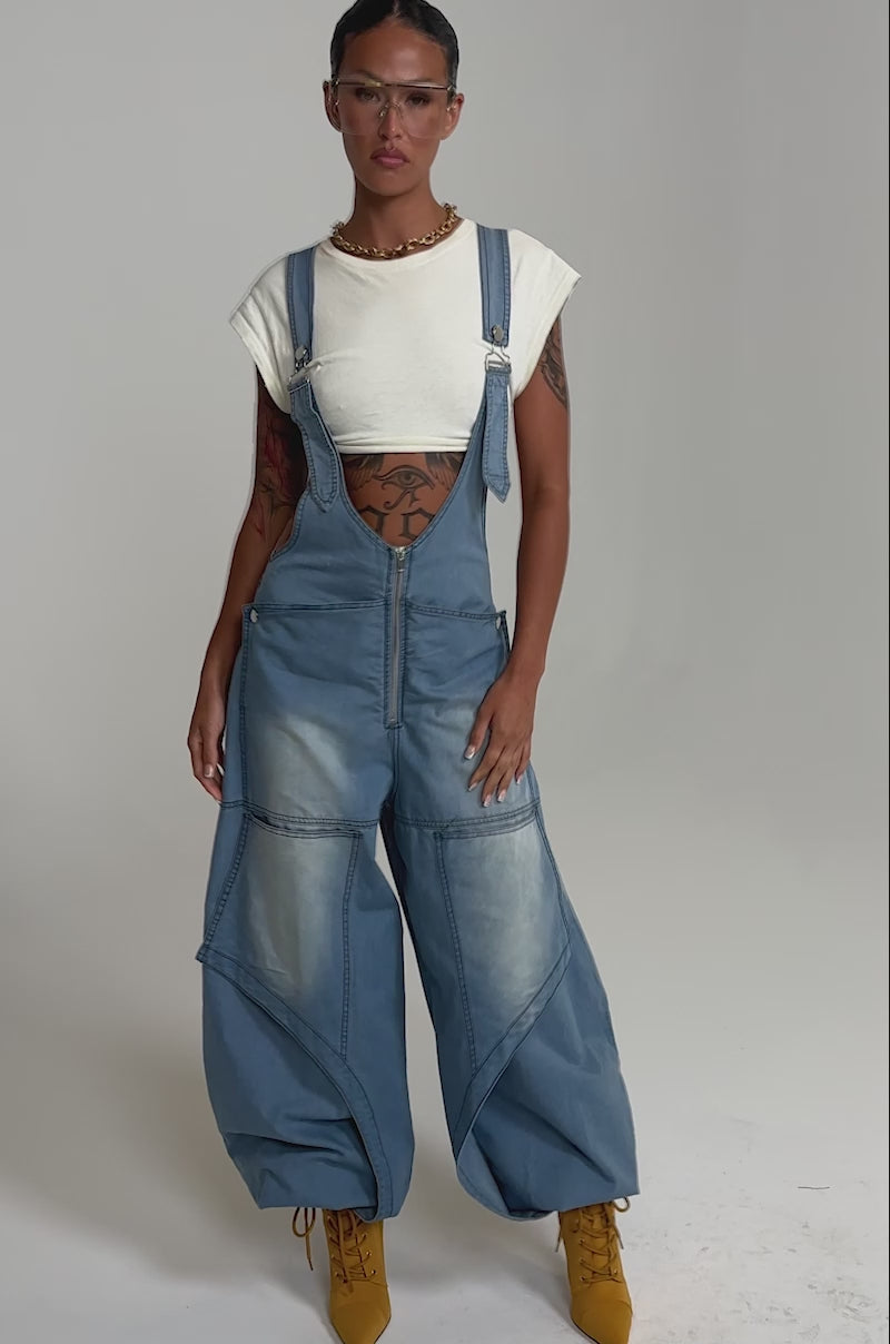 A video of a person wearing the OUT TO PLAY DENIM OVERALL JUMPSUIT with wide legs and a front zipper, styled with a white cropped tee, yellow heeled boots, clear glasses, jewelry, tattoos, and a sleek hairstyle.