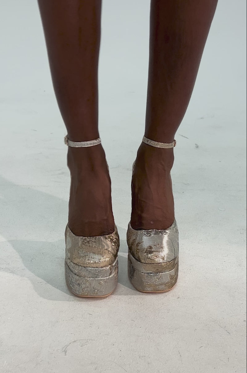 Video of woman modeling the AZALEA WANG ZIPPHORA GOLD MULTI BROCADE PLATFORM PUMP with chunky block heels, ankle straps, and a shimmering gold and silver brocade pattern