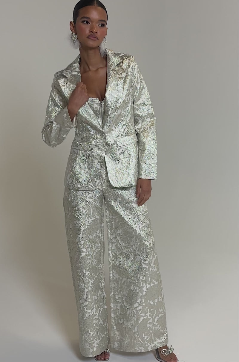 Video of woman modeling the COCO BROCADE BLAZER, a shimmering textured silver longline pantsuit with a deep neckline. She completes the look with slicked-back hair and statement earrings.