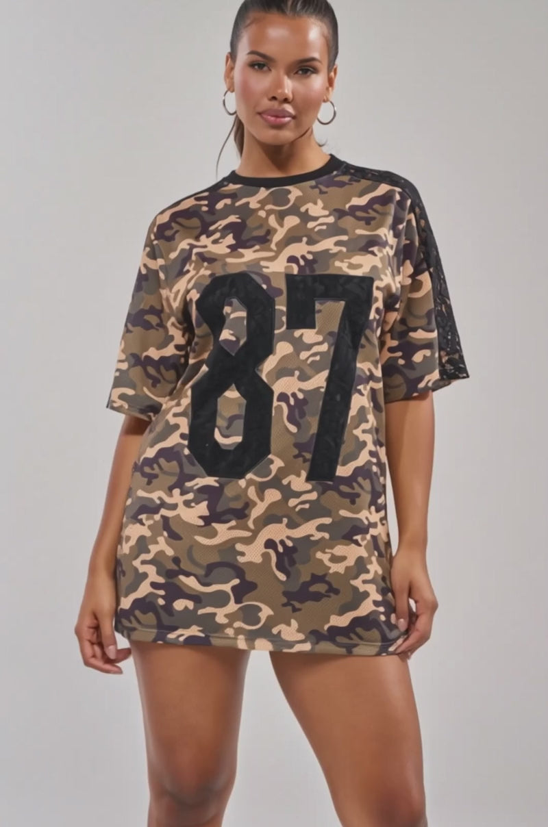 Video of woman modeling the POPPIN POPPYS Lace Jersey Mini Dress in camo, featuring a sporty-chic loose fit, black lace on one sleeve, and a bold black "87" on the front.