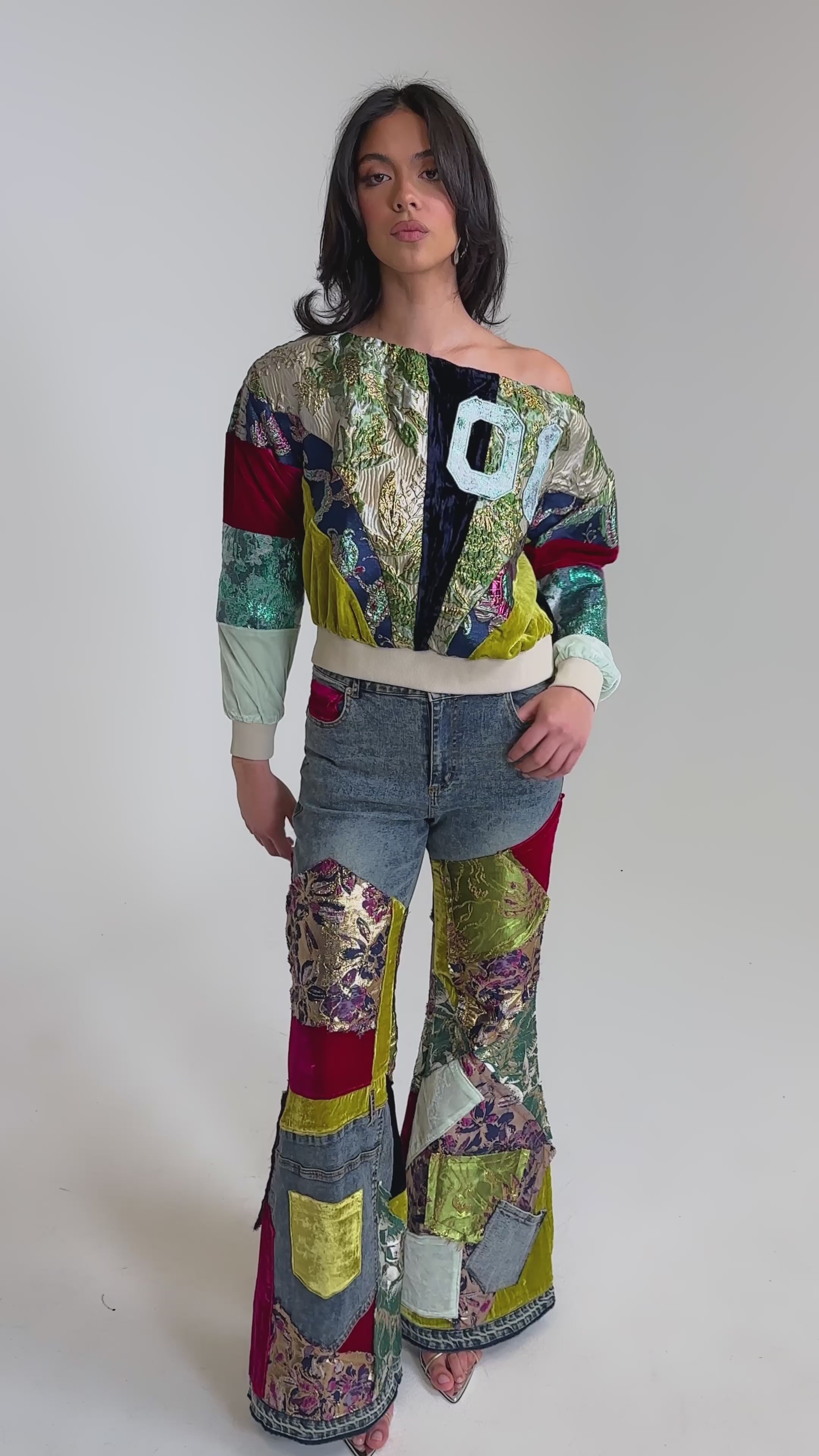 A video of a woman wearing the MIXED EMOTIONS BROCADE PATCHWORK FLARE DENIM—high-rise flared jeans and a matching patchwork jacket in assorted colors, velvet, and floral fabrics—posed against a plain light gray background.