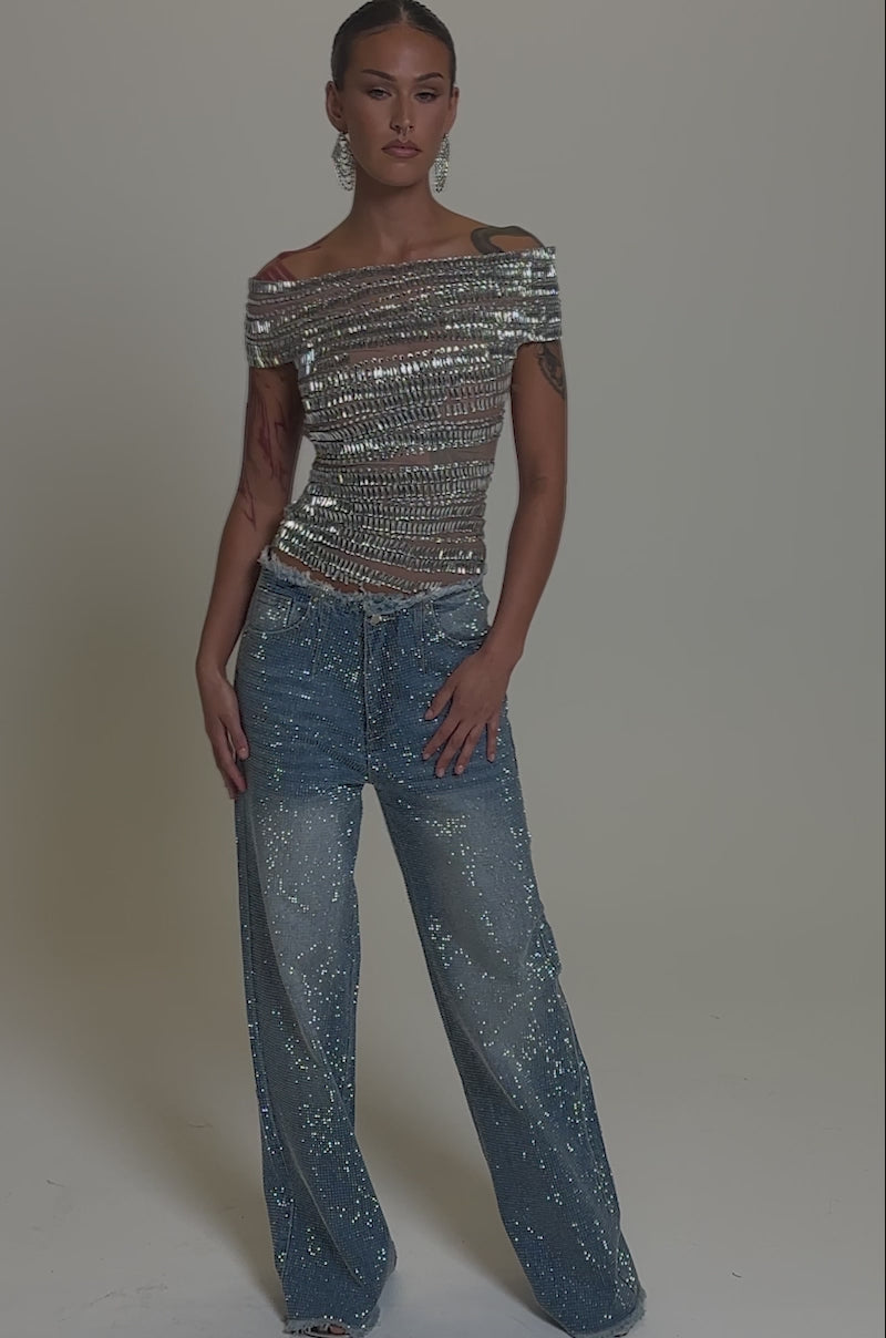 Video of woman modeling the SHINE LIKE A DIAMOND RHINESTONE MESH BLOUSE with silver horizontal embellishments, paired with blue embellished jeans. She wears large dangling earrings, showcasing tattoos on her arms against a neutral background.