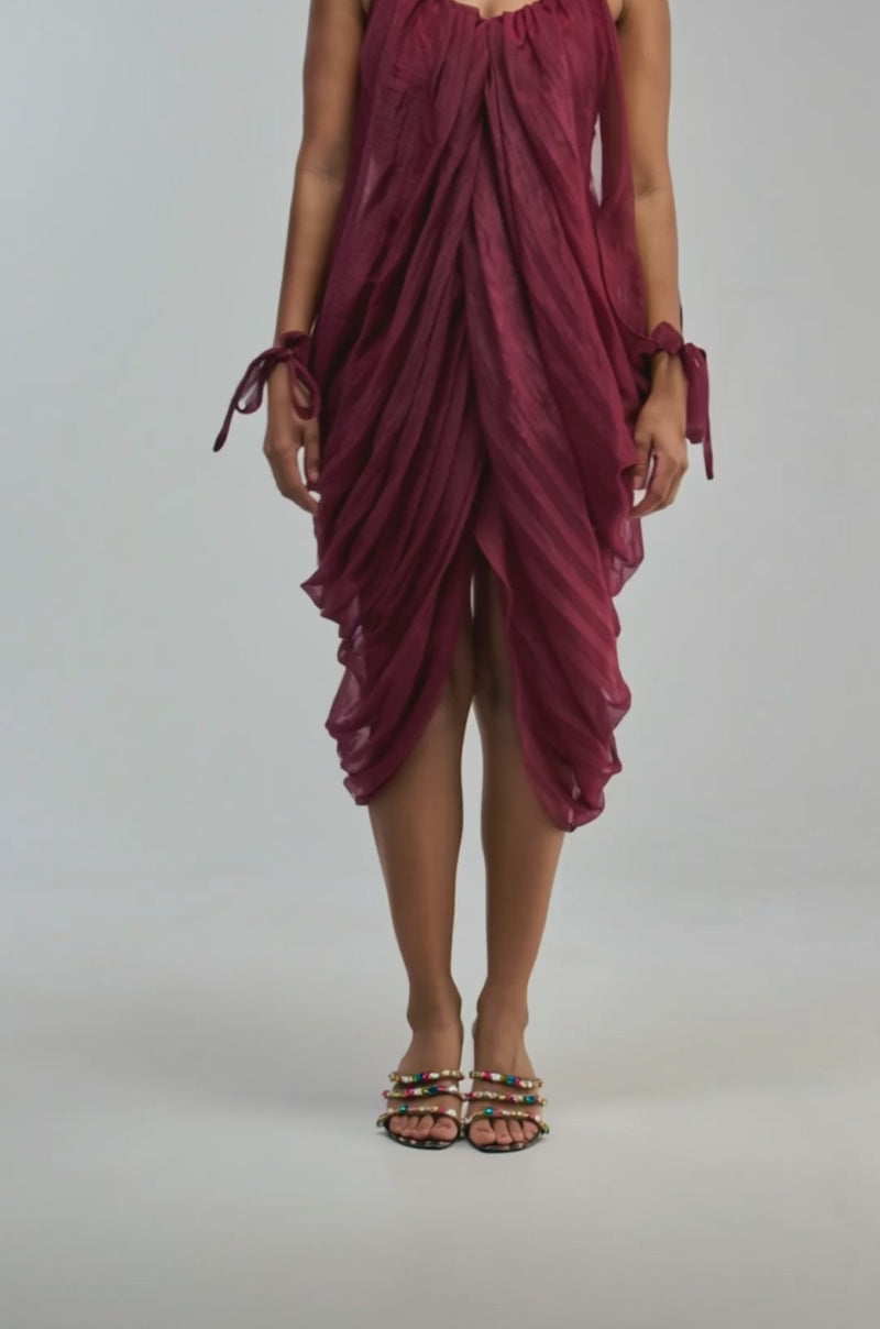 Video of a woman modeling the VIVA LA VIDA MESH DRESS IN BURGUNDY—a sleeveless chiffon mini dress with thin straps, side ties, and elegant draping.