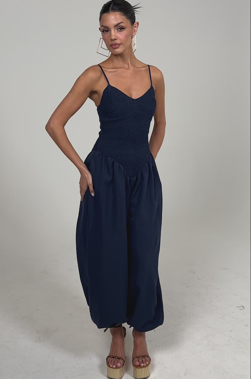 Video of woman modeling the TRENDSETTER BALLOON PANT JUMPSUIT featuring a smocked navy bodice, thin straps, and loose, gathered ankle pants. Paired with platform wedge sandals, it's perfect for an elegant evening look against a simple backdrop.