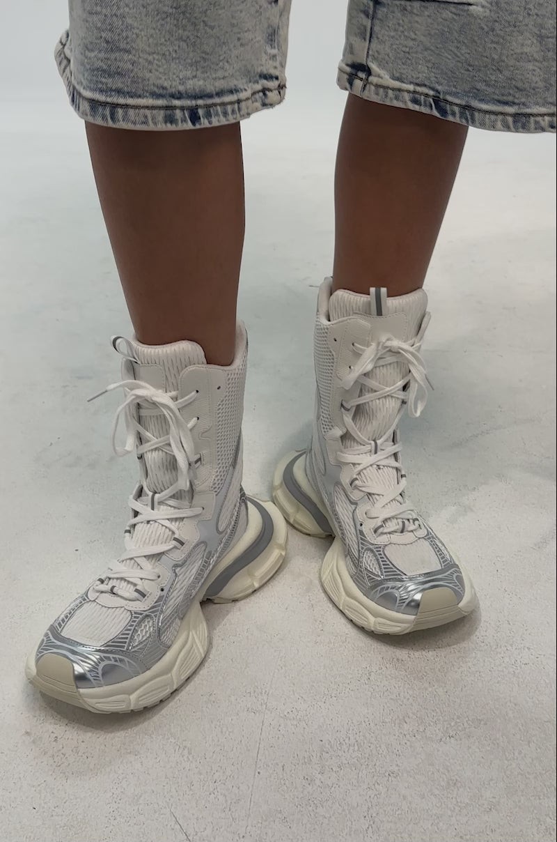 A video of a model wearing the AZALEA WANG YVONNE WHITE LACE UP SNEAKER. The sneaker is a metallic high-top with a chunky cream sole, white mesh upper, shiny silver accents, white laces, and a heel pull tab for a sporty look.