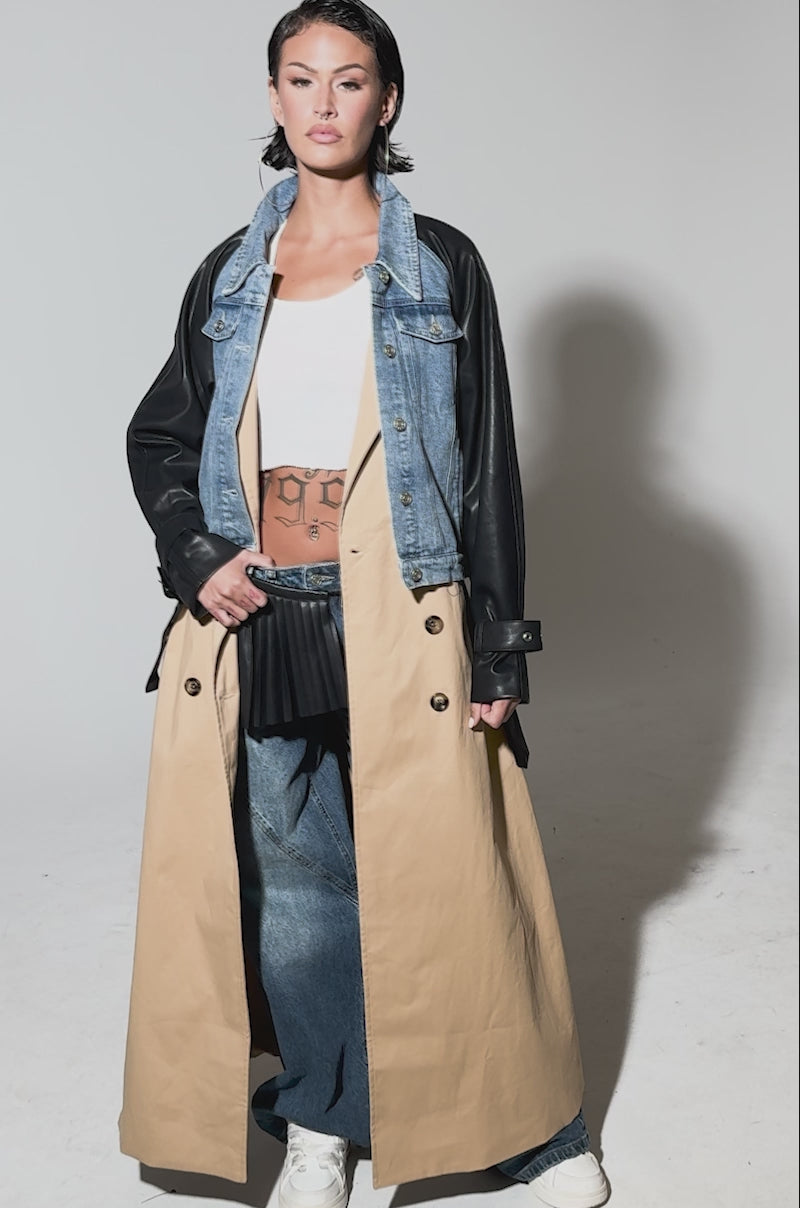Video of woman modeling CITY SLICKER MIXED MEDIA TRENCH with black leather sleeves, a wide belt, beige double-breasted skirt detail, blue jeans, white shoes, and large hoop earrings.