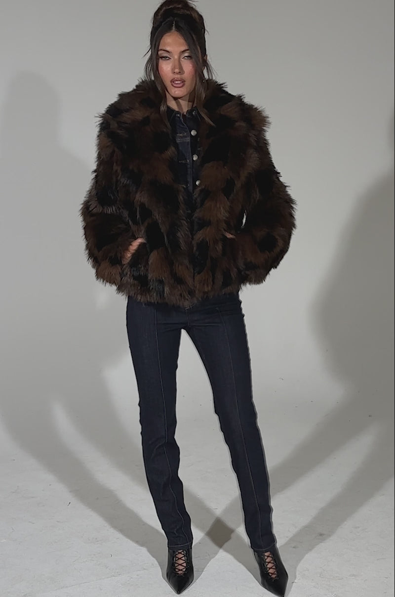 A video of a woman standing confidently against a light blue background, wearing the SHORTIE FUR COAT IN BROWN MULTI, paired with dark denim jeans and black heeled lace-up boots.