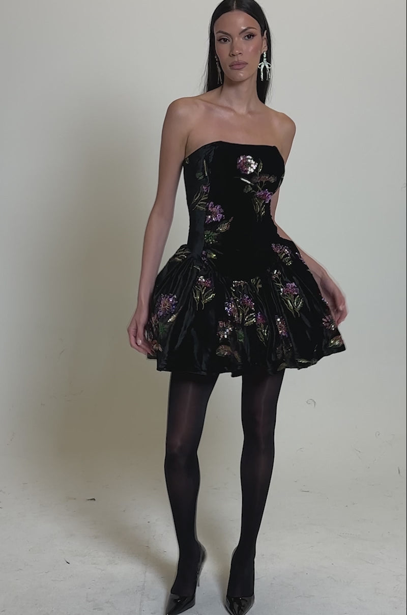 A video of a woman in the LATE NIGHT GARDEN PARTY VELVET MINI DRESS, a strapless black mini with floral embroidery and a bubble skirt. She pairs it with black tights, heels, straight dark hair, and stands before a plain gray background.
