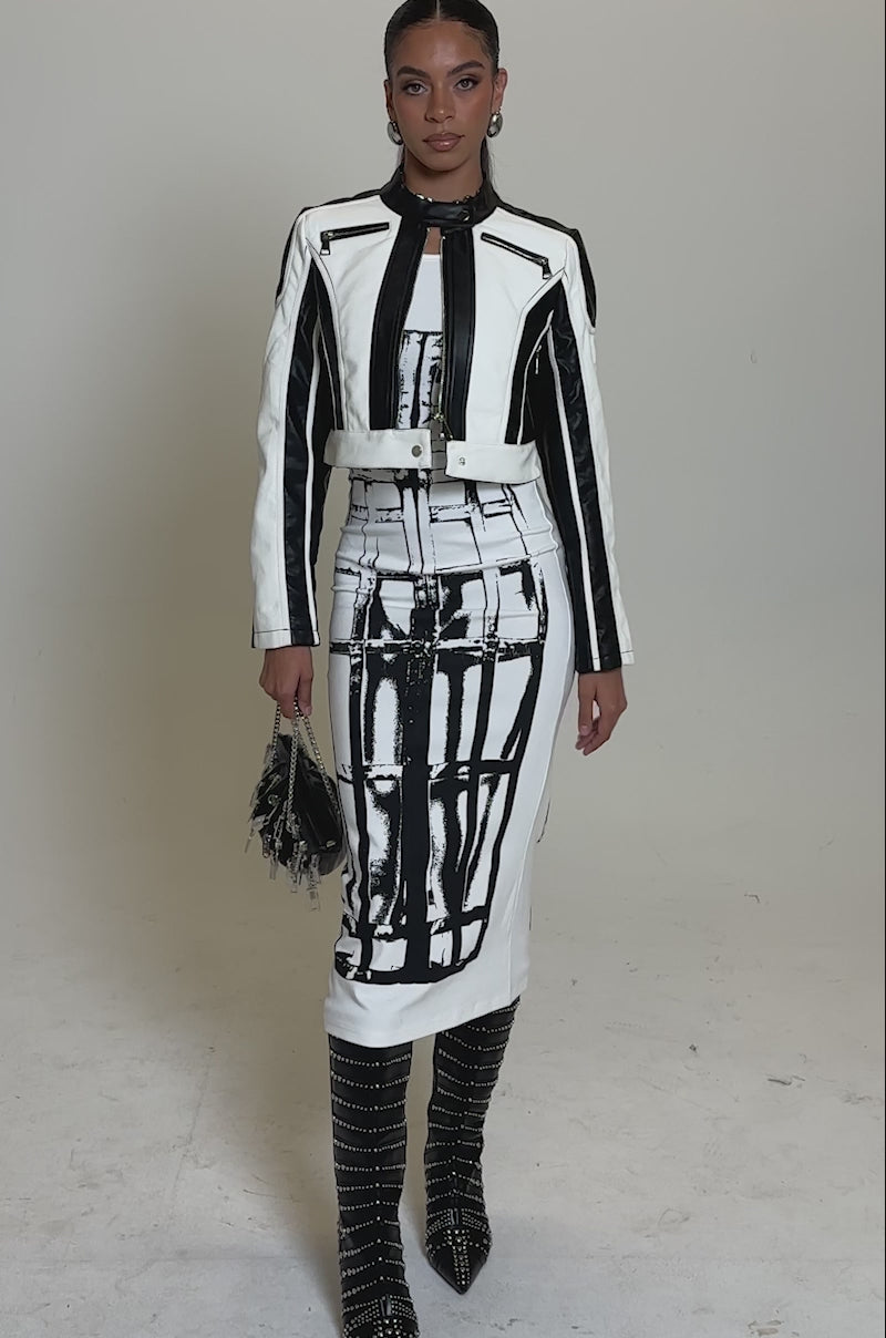 A video of a woman wearing the BETTER LUCK CROPPED MOTO IN BLACK WHITE jacket, a matching abstract-patterned knee-length dress, studded knee-high boots, and carrying a small black purse with metal details.
