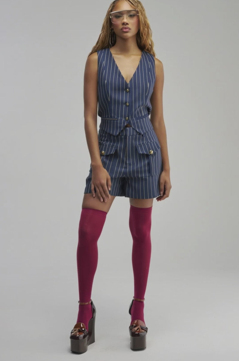 Video of a woman modeling the  SUNDAY PINSTRIPE VEST as part of a stylish three-piece suit, a person with long, light brown braided hair and clear glasses stands against a plain gray background.