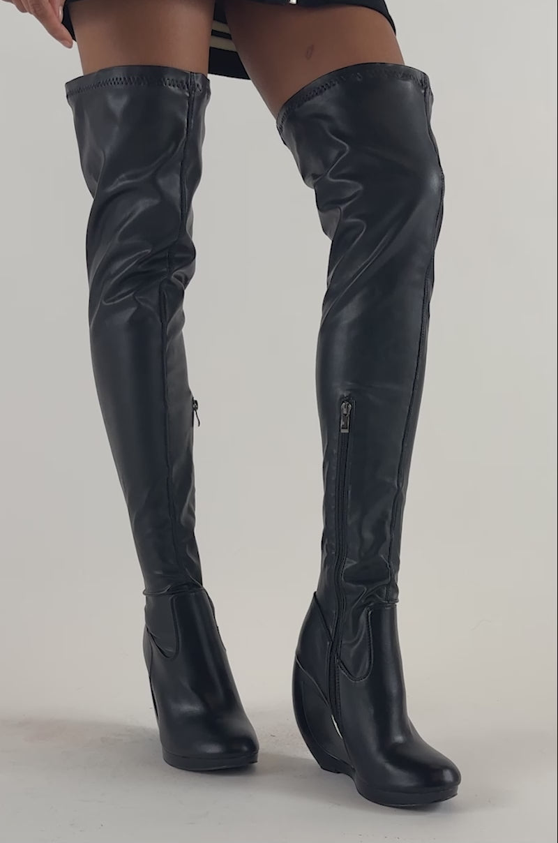 Video of a woman modeling the AZALEA WANG CARSON BLACK 4 WAY STRETCH PU OVER THE KNEE OPEN WEDGE BOOT, which features a unique cutout heel.