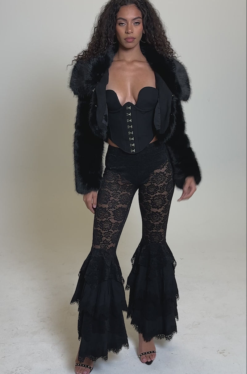 Video of woman modeling the CADENCE FAUX FUR IN BLACK over a black corset top and lace pants, standing against a plain light gray backdrop.