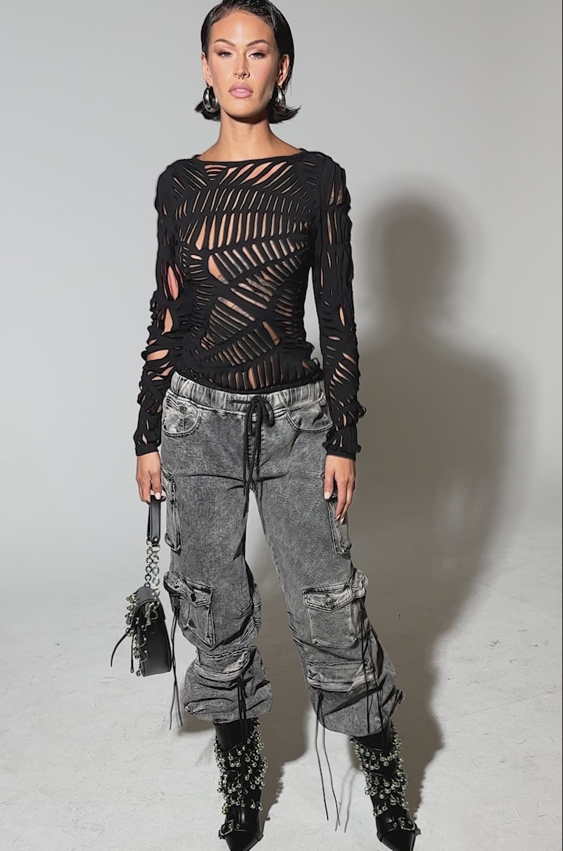 Video of woman modeling the CARGO FOR DAYS DENIM JOGGER—distressed black streetwear cargo pants with zippers and drawstrings—paired with a ripped black long-sleeve top, chain-detailed black boots, and a small black bag with metallic accents.