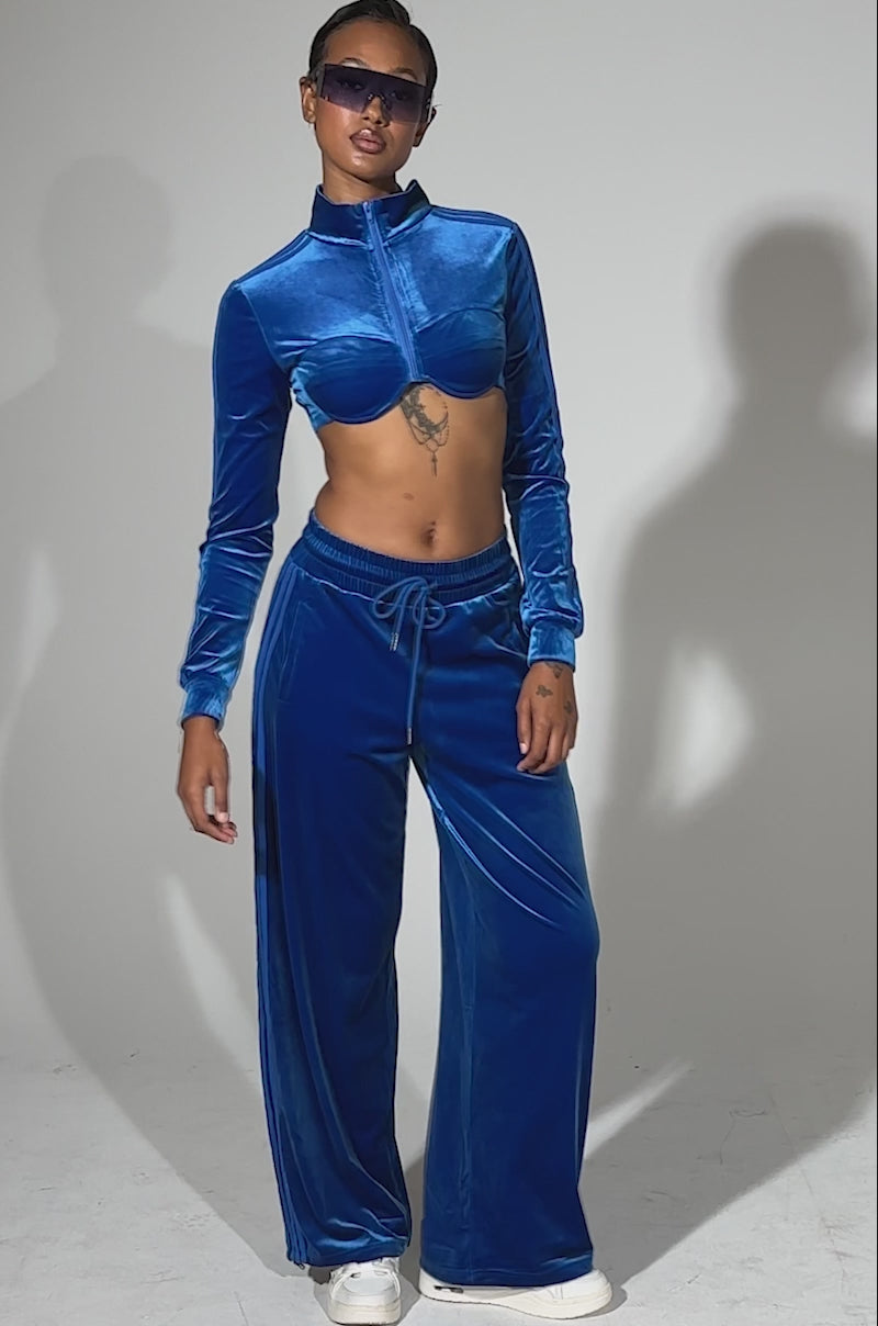 A video of a person standing confidently in the ELECTRIC BLUES CROP ZIP UP SWEATSHIRT and matching sapphire-blue velour wide-leg pants, paired with white sneakers and sunglasses. They have short hair plus visible tattoos on their torso and hand.