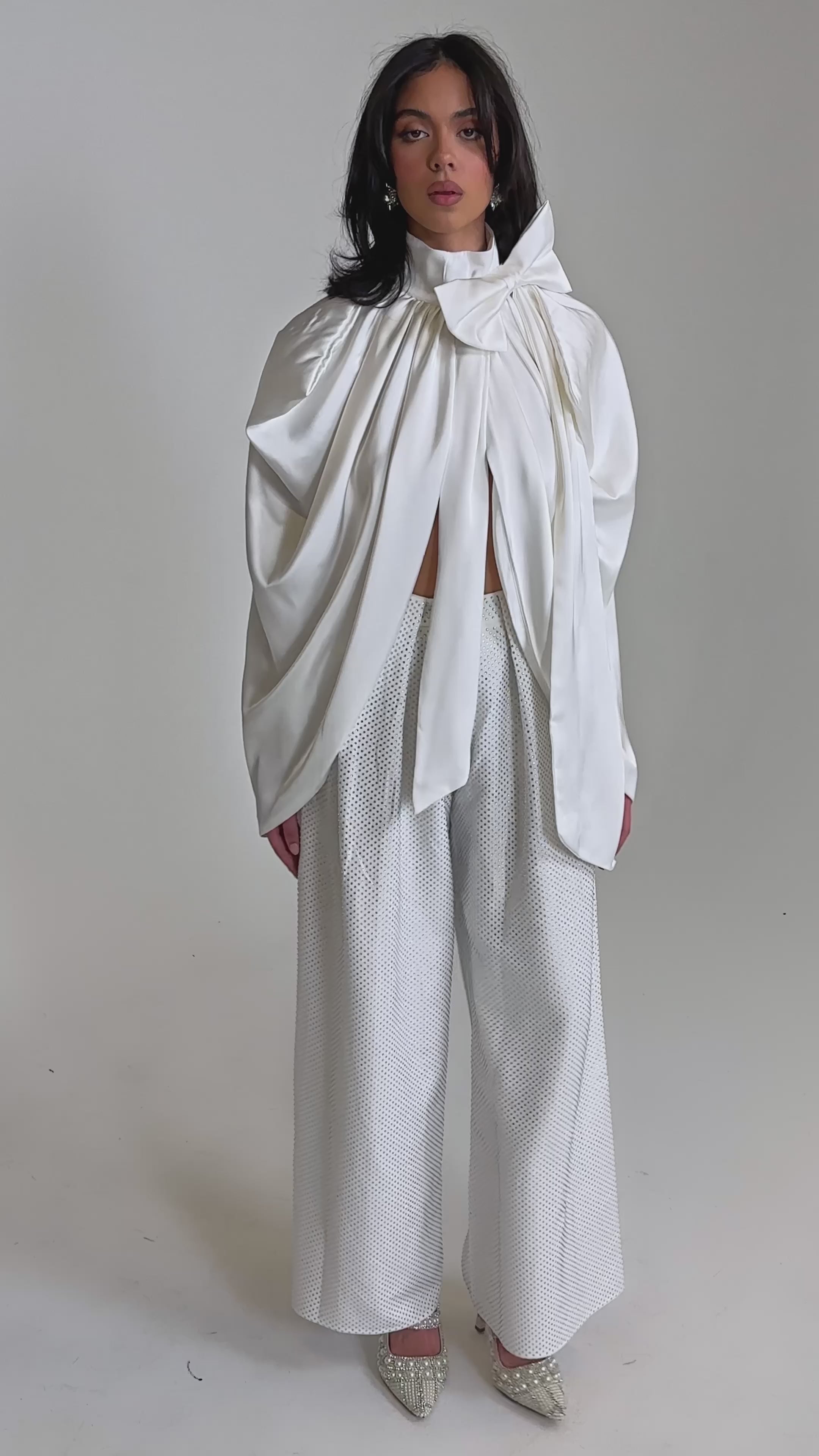 A video of a woman in the WRAPPED IN LOVE BLOUSE, featuring a dramatic sculptural bow and oversized white satin drapes, paired with textured wide-leg trousers and pointed-toe shoes against a plain light background.