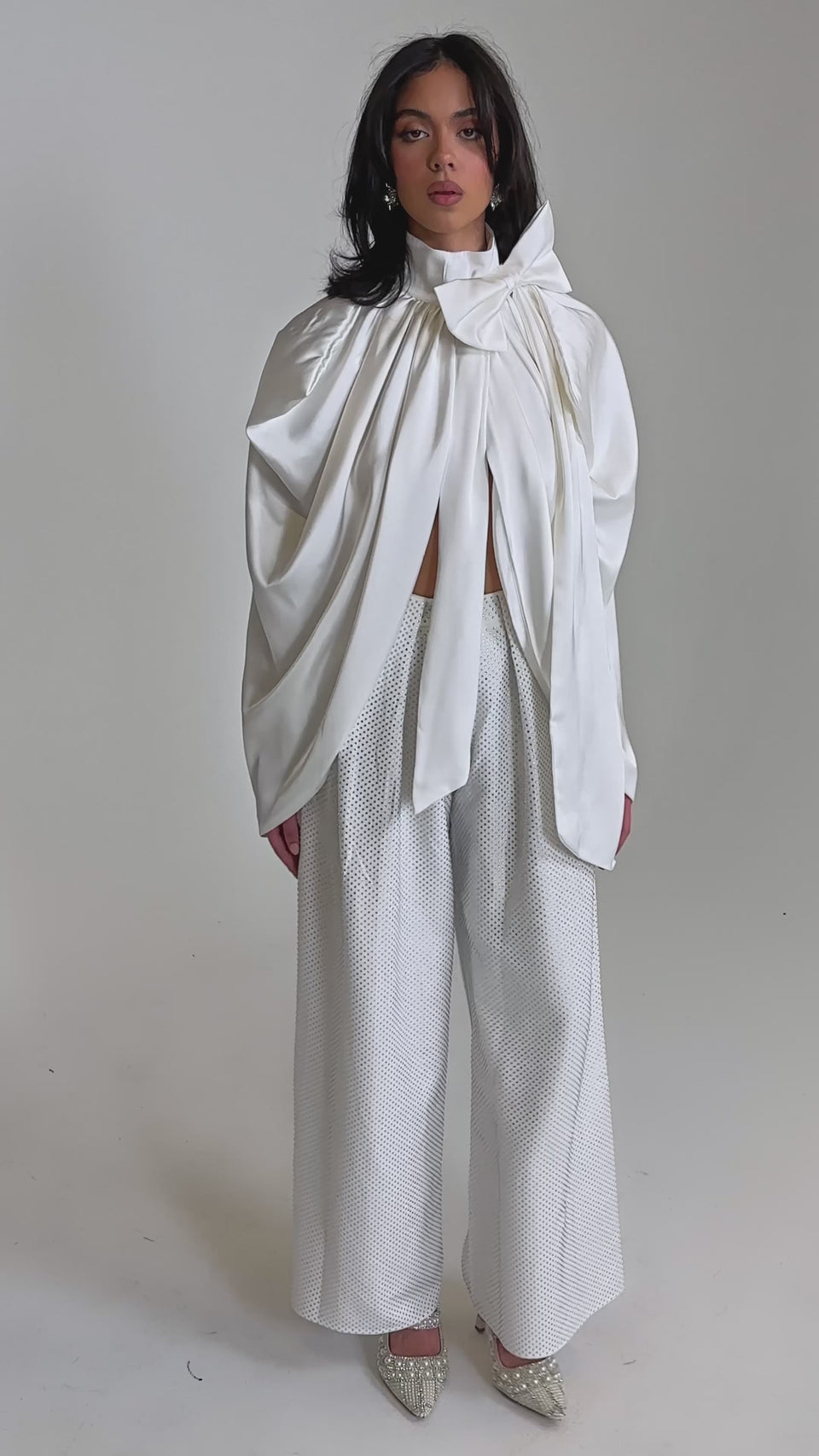 A video of a woman in the WRAPPED IN LOVE BLOUSE, featuring a dramatic sculptural bow and oversized white satin drapes, paired with textured wide-leg trousers and pointed-toe shoes against a plain light background.