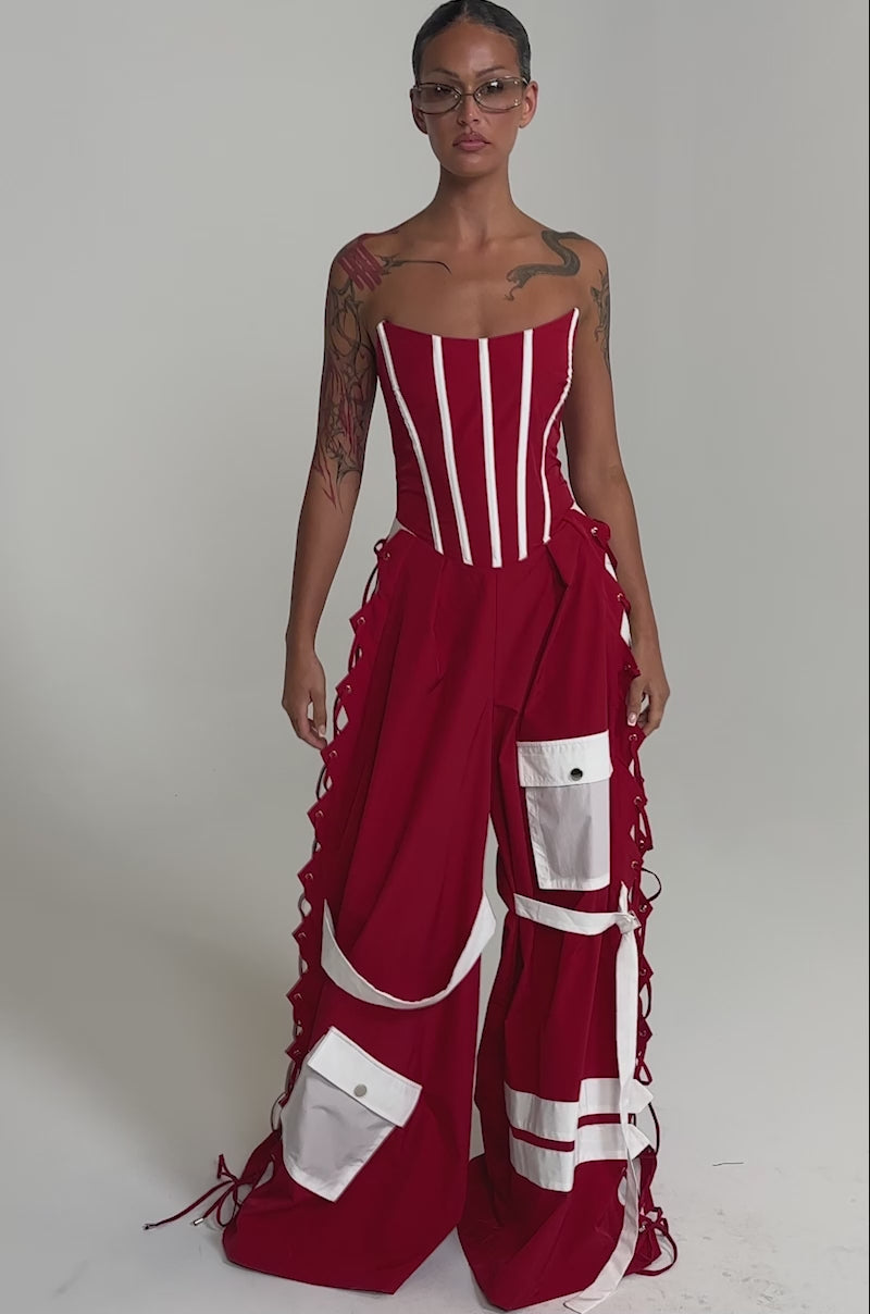 A video of a person in the SPORTY VIBE CORSET JUMPSUIT, featuring a bold red corset-style top, wide-legged pants with white stripes, large pockets, lace-up sides, and glasses.