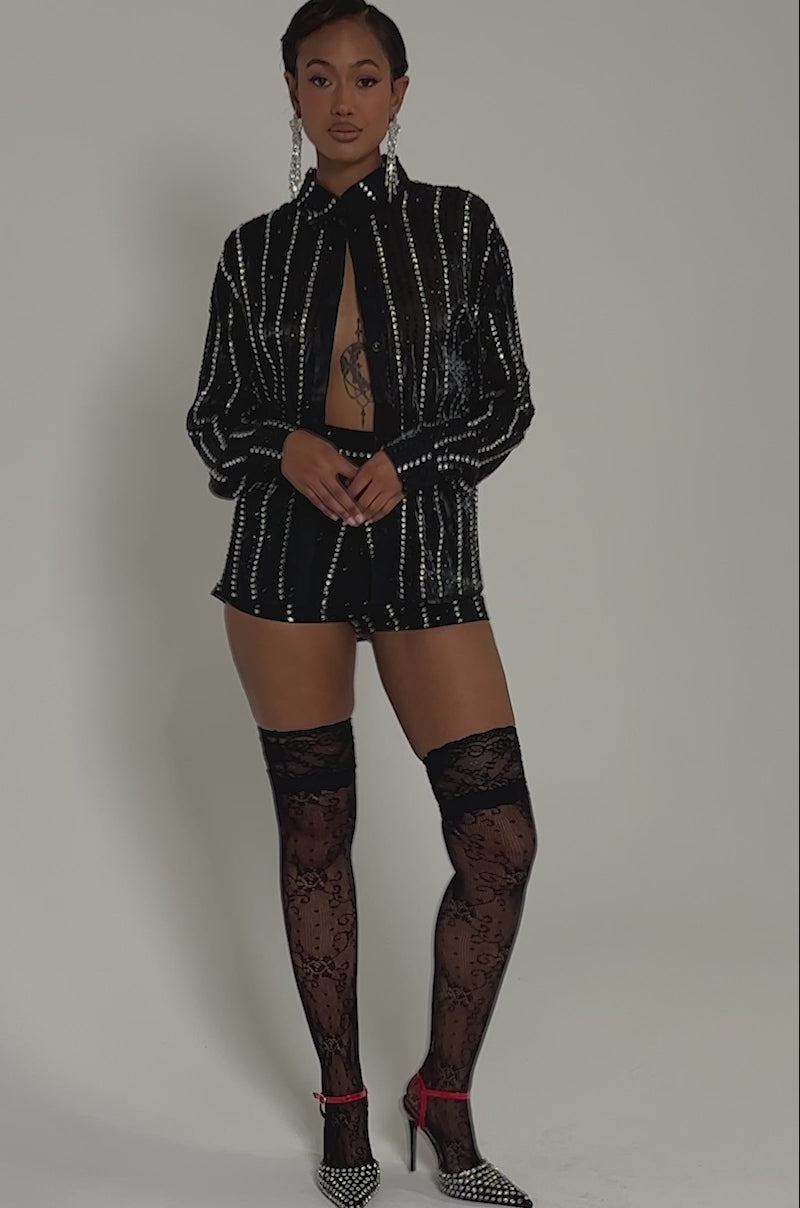 Video of woman modeling the DANCE THE NIGHT AWAY SHORT, a black, sparkly, long-sleeved romper with vertical stripes, paired with black lace thigh-high stockings, studded high heels, and statement earrings against a plain background.