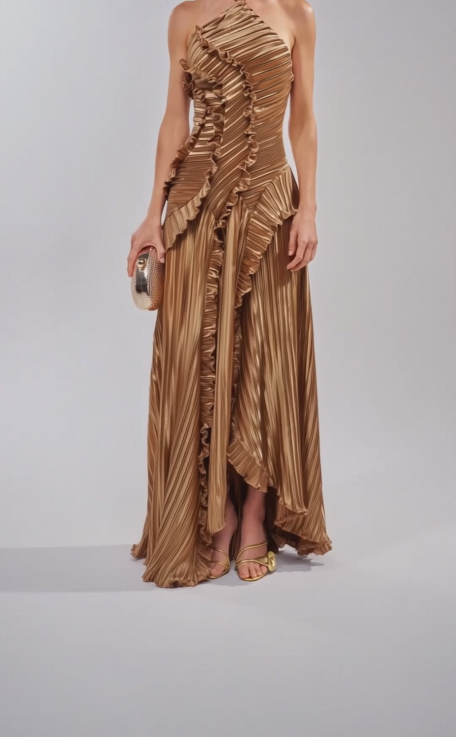 A video of a woman in the CELESTE RUFFLE MAXI DRESS IN BRONZE—a pleated, one-shoulder gown with sculptural ruffle detailing—paired with a gold clutch, earrings, and strappy sandals.