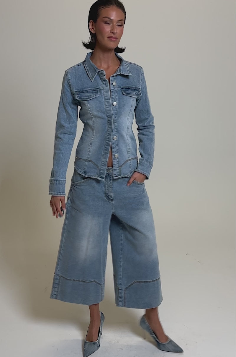 A video of a woman in the ROCK MY WORLD DENIM BUTTON DOWN with matching wide-leg cropped jeans and blue heels against a plain, light grey background.