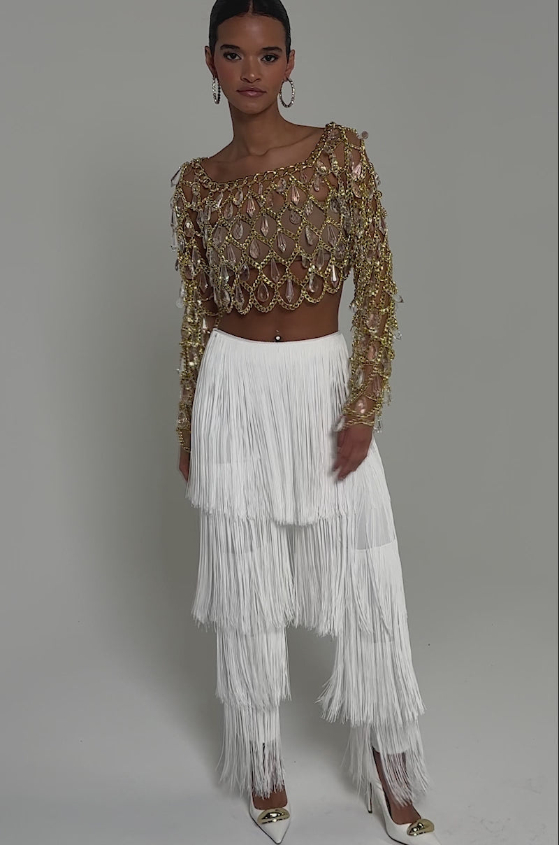 Video of woman modeling the OH SO FRINGE PANTS IN WHITE, styled with a gold embellished long-sleeve crop top, hoop earrings, gold pointed heels, and her hair pulled back.
