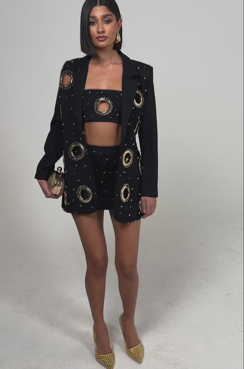 A video of a woman wearing the SHOW STOPPER MINI SKIRT with large gold ring cutouts, paired with a matching black crop top, blazer, gold heels, hoop earrings, and a small gold clutch.