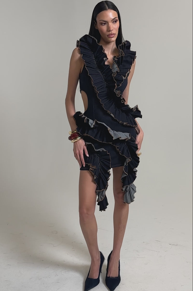 A video of a woman wearing the SAY IT AGAIN RUFFLED DENIM MINI DRESS IN DARK BLUE DENIM, featuring sleeveless cutouts and bold ruffle details. 