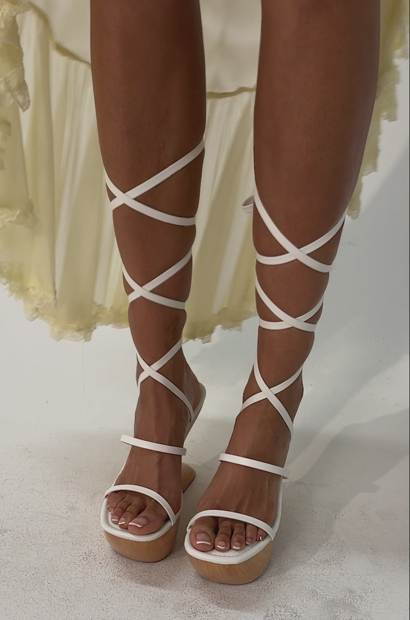Video of woman modeling the AZALEA WANG ONNI WHITE STRAPPY UPPER WOOD WEDGE sandals, featuring sculptural wooden wedge heels and white faux leather straps that lace up her calves.