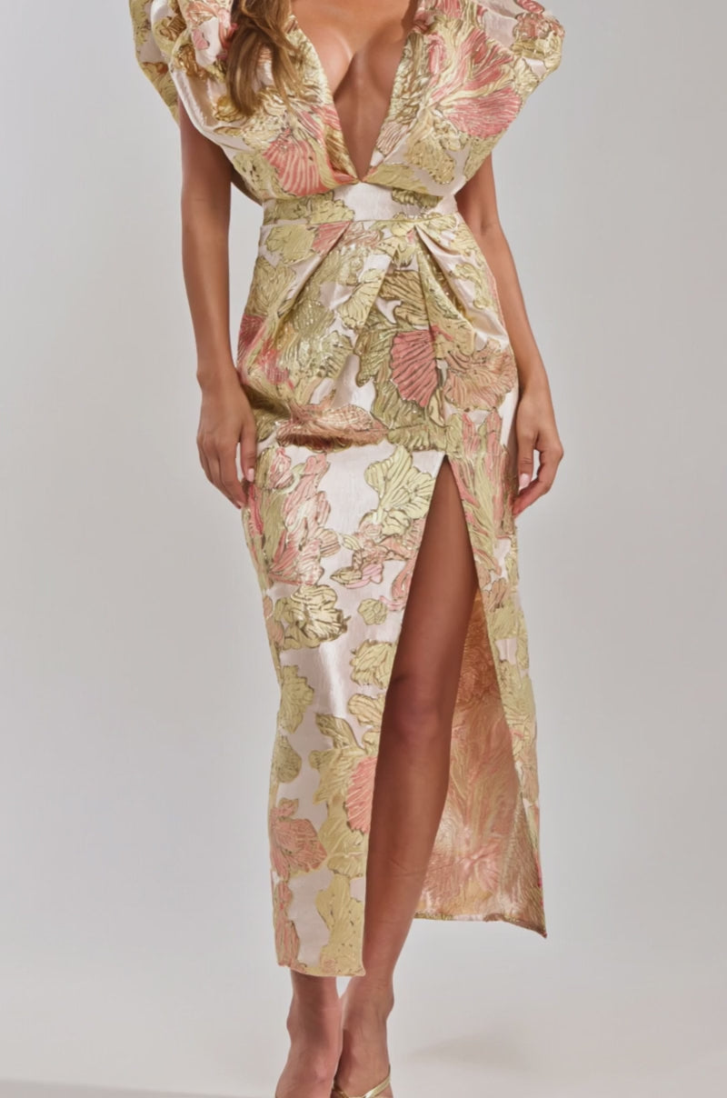 Video of woman modeling the BEST DRESSED BROCADE MAXI DRESS IN PINK, featuring a floral pattern, deep V-neck, structured shoulders, and high slit. She styles it with statement earrings and gold heels, her long hair flowing against a plain background.