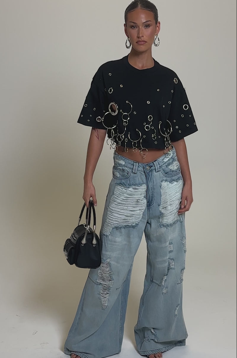 Video of woman modeling the RING AROUND ME CROPPED TEE with silver grommet and ring details, paired with light blue distressed jeans, hoop earrings, slicked-back hair, and carries a black handbag by her side.