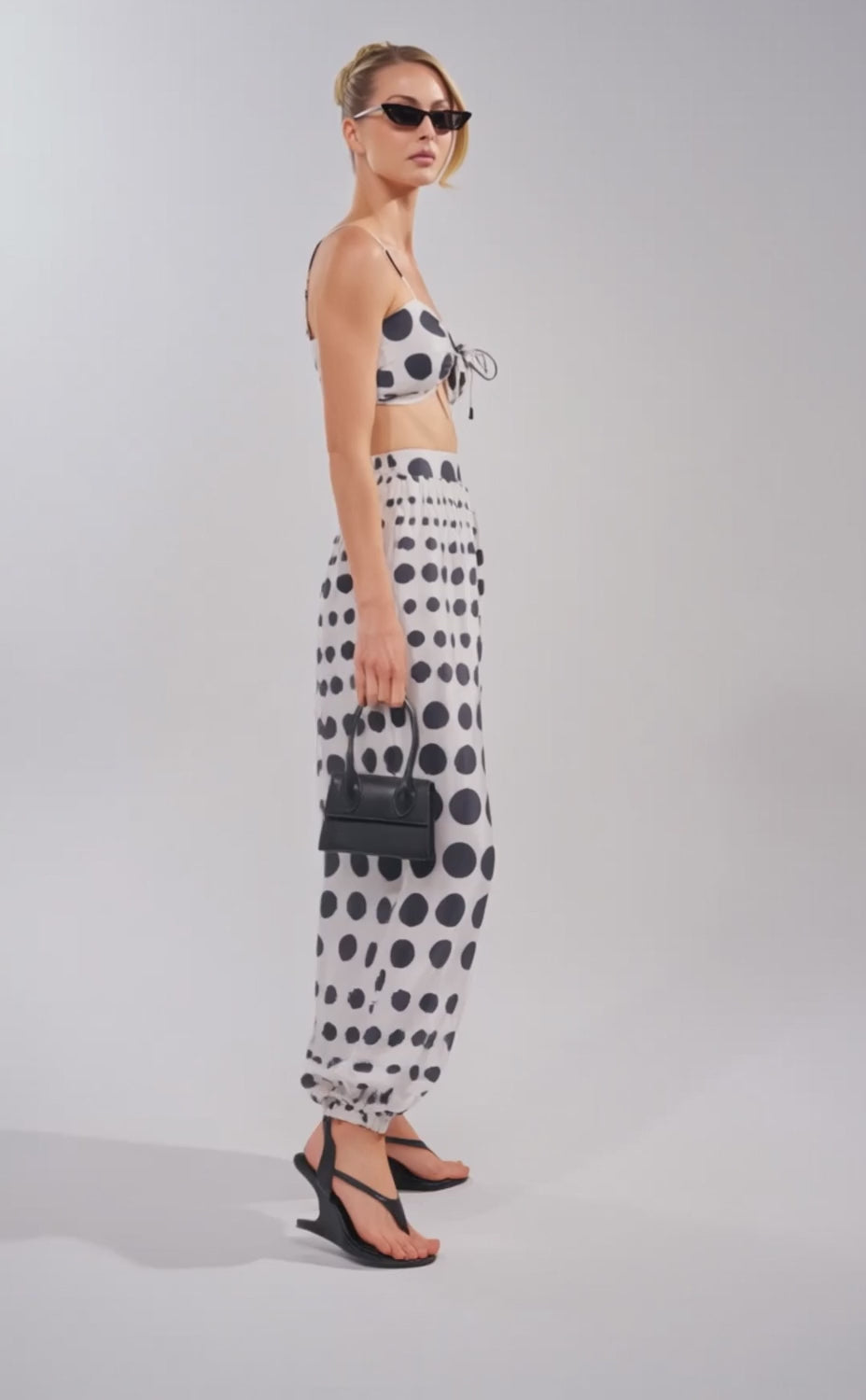 A video of a woman wearing the POLKA DOT BIKINI GIRL SET—sunglasses, a black and white polka dot bralette with matching high-waisted balloon pants—paired with black sandals and a small black handbag.