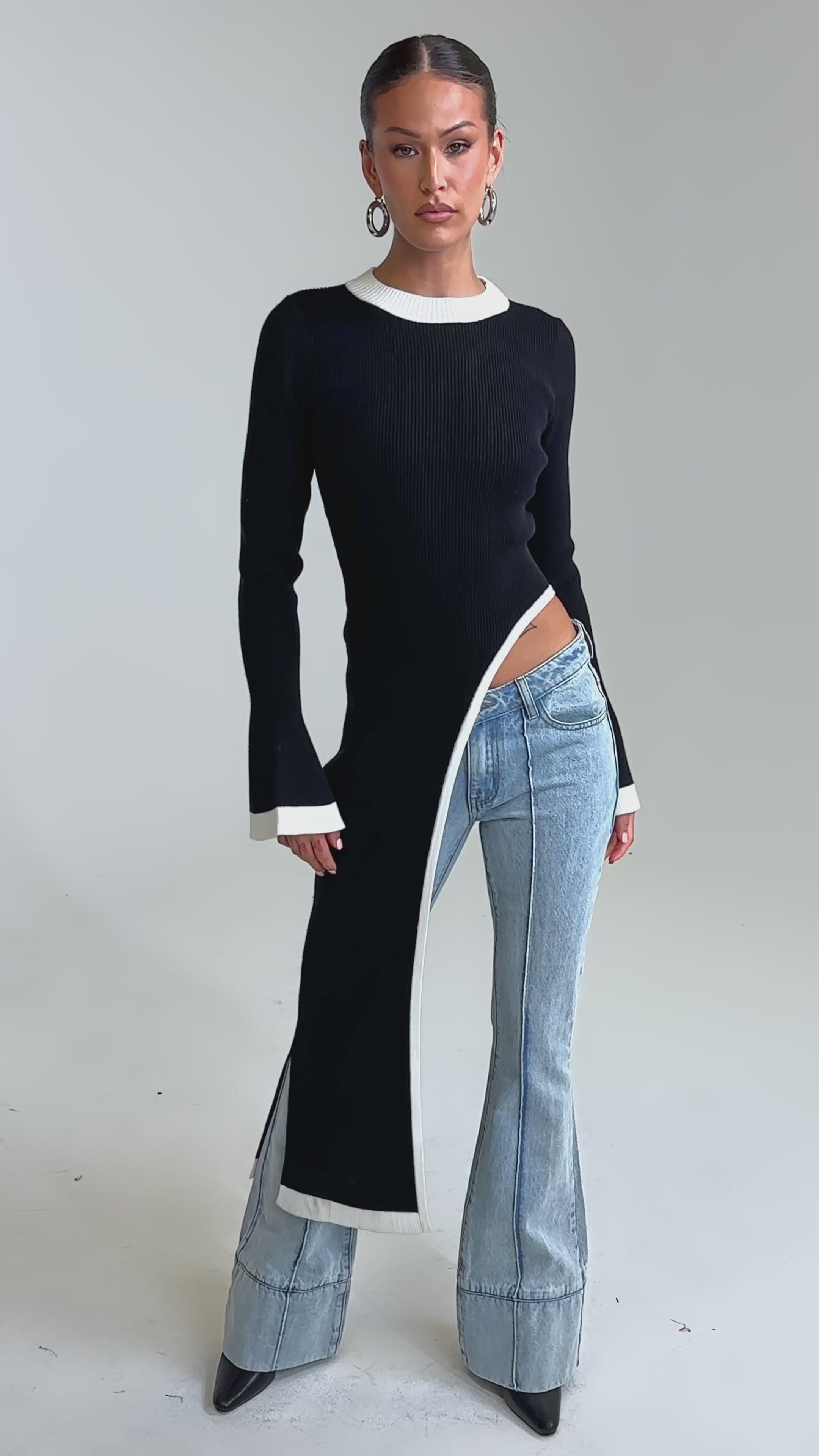 Video of woman modeling the SUPER SOFT EXTENDED SWEATER, a black asymmetrical piece with a white-trimmed hem, long on one side past her knee and short on the other to reveal her hip, paired with light blue flared jeans.