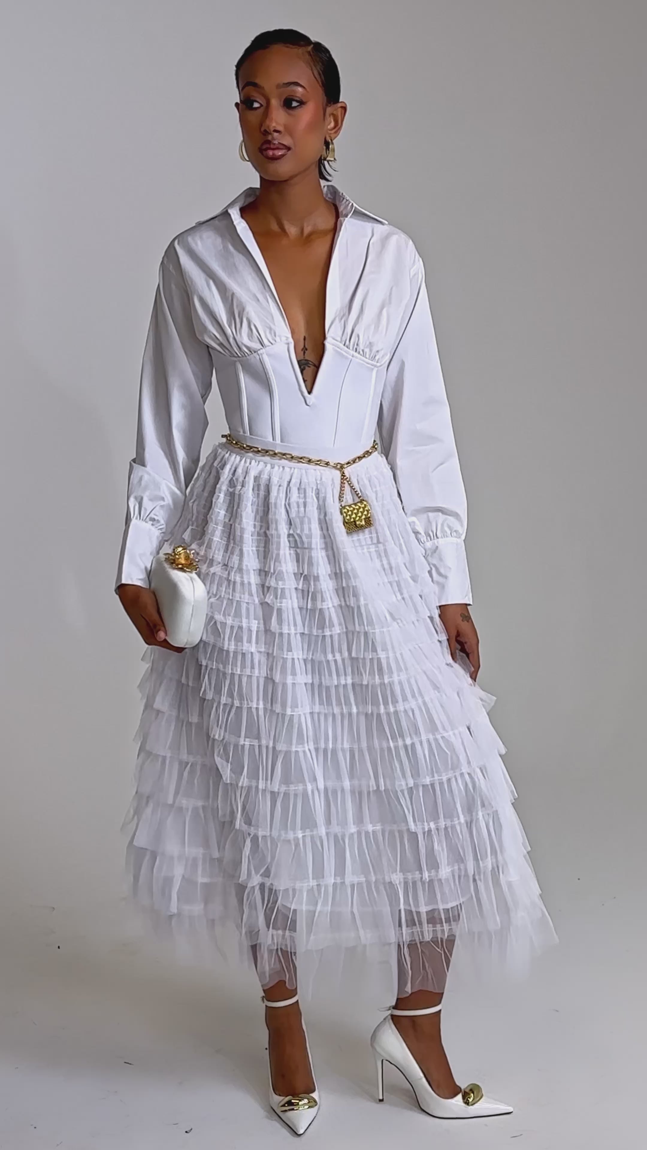 A video of a woman wearing the STARS ALIGNED MESH RUFFLE MAXI SKIRT IN WHITE with a deep V blouse, gold belt, white clutch, gold earrings, and heels, standing against a plain light background.