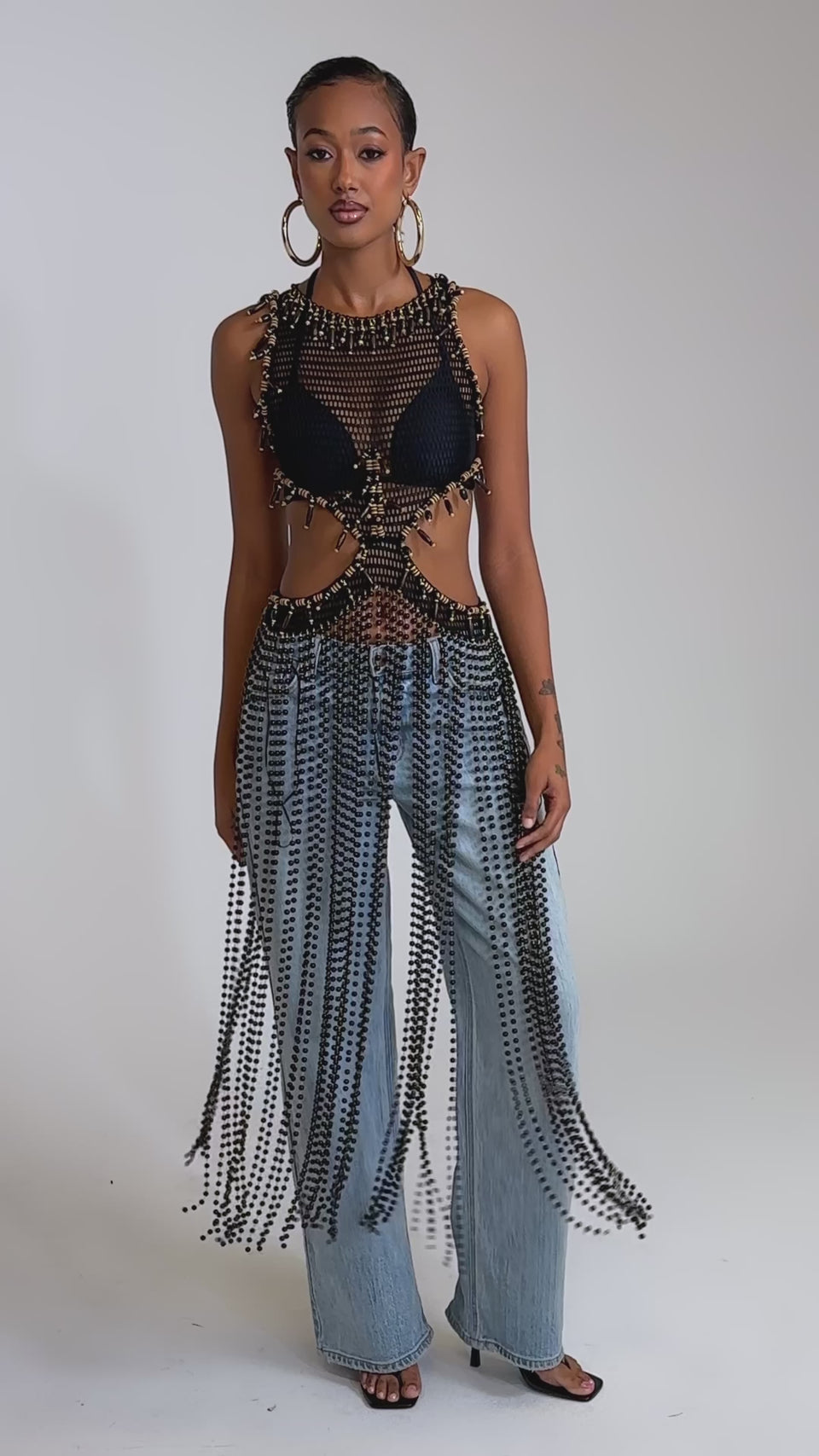 A video of a woman wearing the DRIP DRIP CROCHET BLOUSE over a black bra, light blue wide-leg jeans, large hoop earrings, and black heeled sandals.