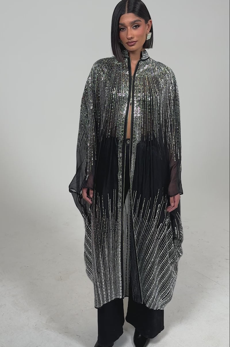 Video of woman modeling the SPARKLE AND SHINE BLOUSE under a long, sheer black cape with silver stripes, paired with black pants and boots. Her hair is styled in a sleek bob.