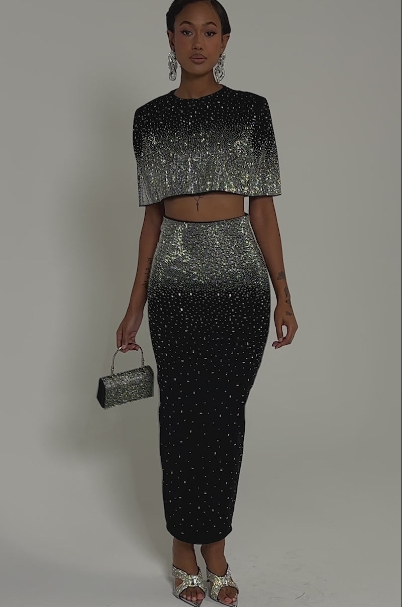 Video of woman modeling the LOVE THIS LIFE BLOUSE, a sparkling, rhinestone-embellished top paired with a sleek black skirt and beaded handbag. Sleek hair and statement earrings complete her perfect evening look against a gray background.