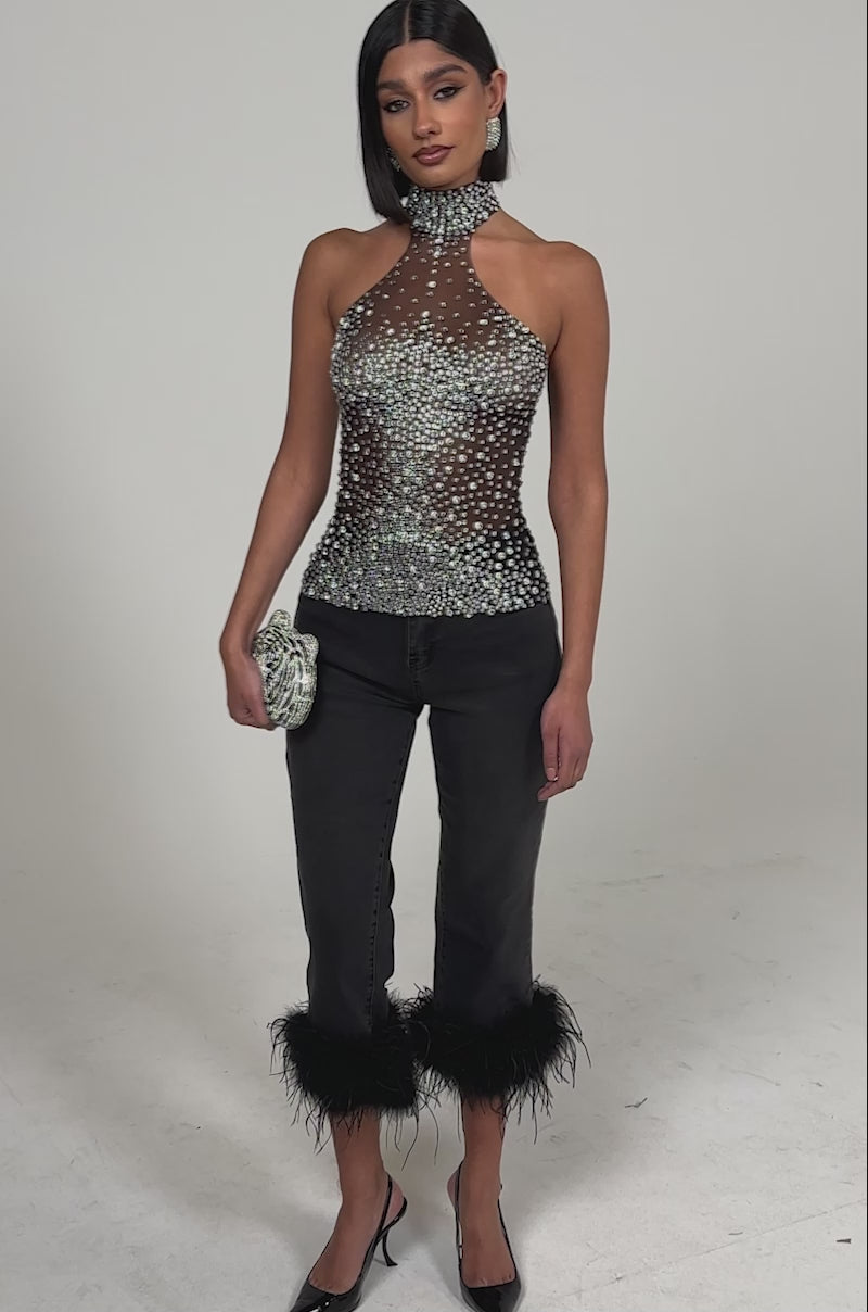 A video of a woman in the ALL YEAR ROUND SHEER RHINESTONE TOP, paired with black feather-hem cropped pants, black pointed heels, and a silver clutch against a plain light background.