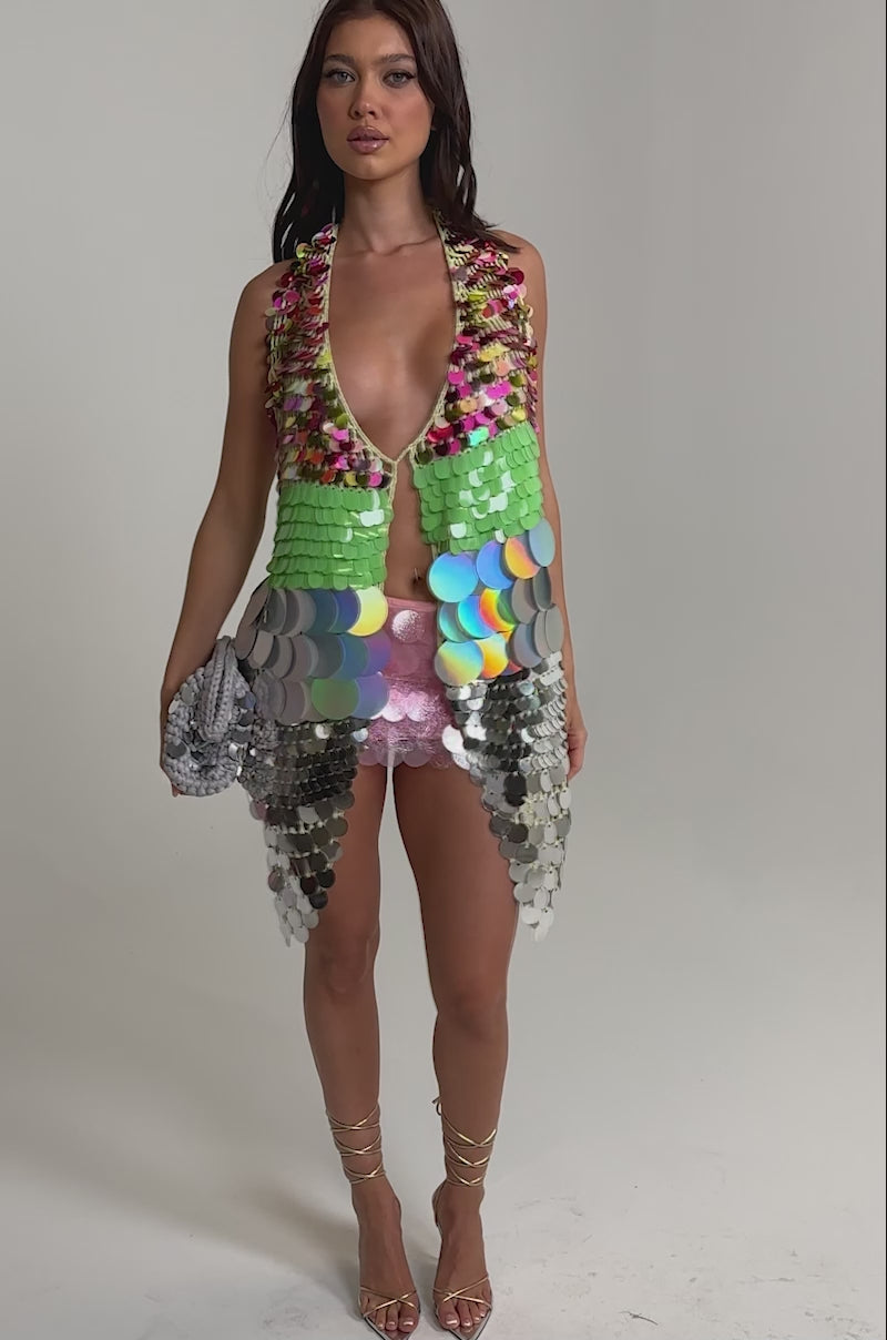 Video of woman modeling the SHAKE IT GIRL CROCHET SEQUIN TOP—a colorful green, gold, and silver festival vest—paired with a pink sequined skirt and a silver clutch, posing against a plain light backdrop.