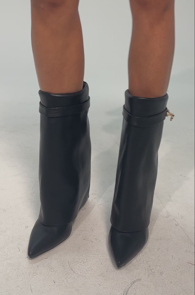 A video of a model wearing the AZALEA WANG LOUISA BLACK PU WEDGE BOOTIE. It is a black faux leather bootie with a wide, angular foldover shaft and a small gold buckle near the top.