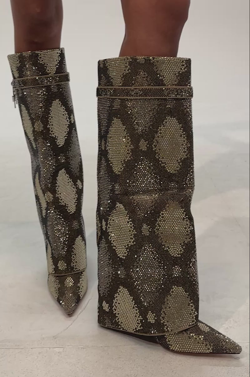 Video of woman modeling the AZALEA WANG EVO KHAKI SNAKE SPARKLE WEDGE BOOT featuring knee-high construction adorned with gold and olive rhinestone embellishments in a snake-inspired pattern, a pointed toe, distinctive small padlock detail against a light gray background.