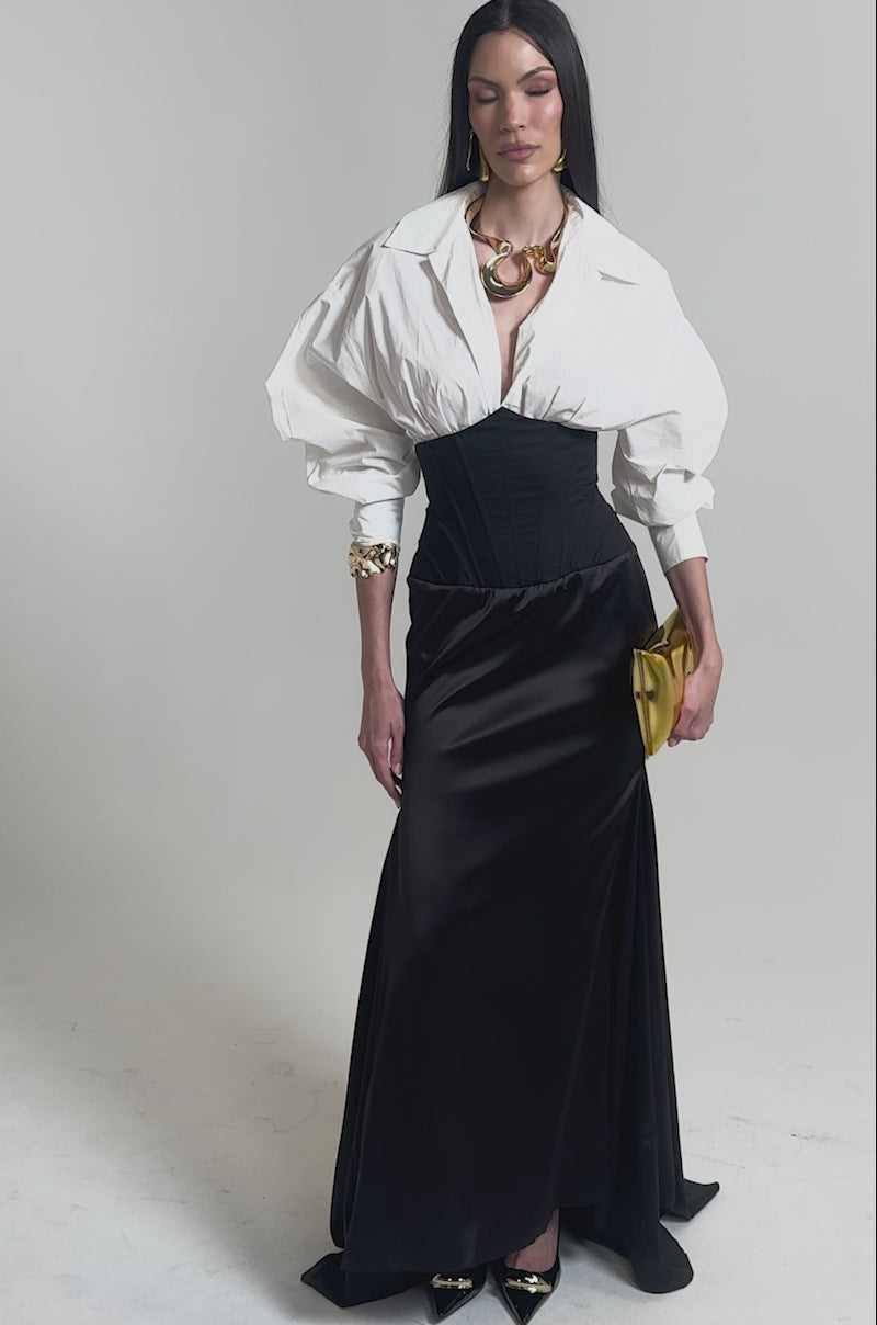 A video of a woman in the STAR OF THE NIGHT HIGH WAISTED CORSET MAXI SKIRT, styled with a white blouse, gold jewelry, a gold clutch, and black pointed heels against a plain light background.