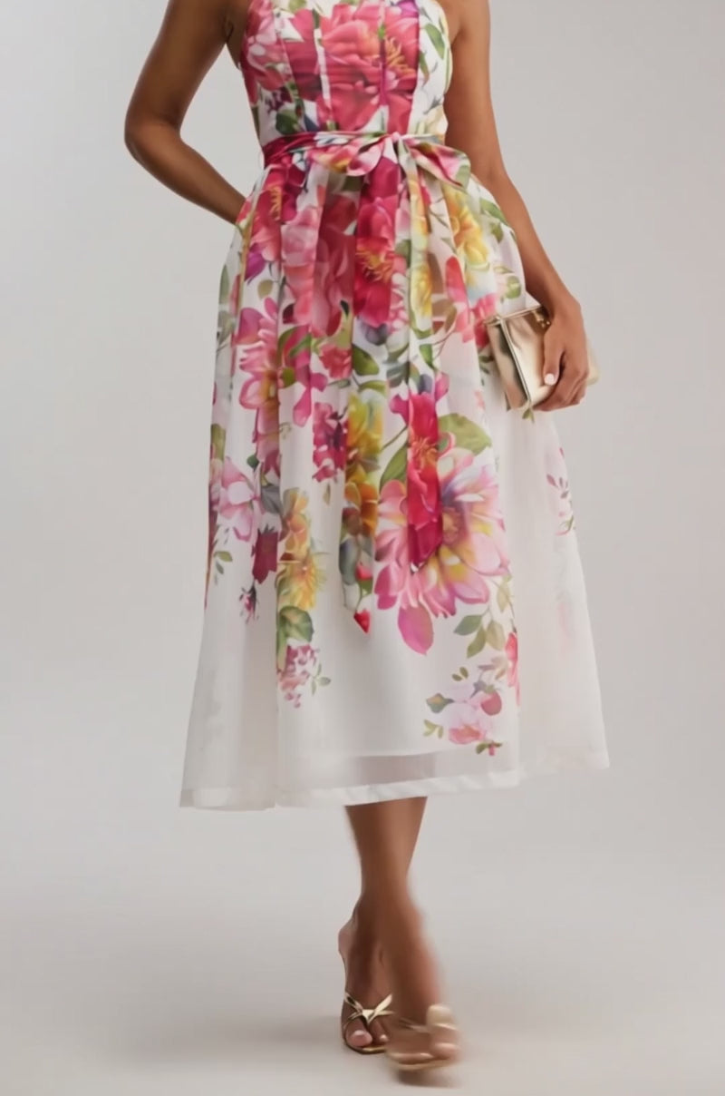 Video of woman modeling the GARDEN PARTY FLORAL CHIFFON MIDI DRESS with a fitted bodice and pink ribbon belt, paired with a gold clutch, gold heels, and a sleek bun—an elegant look for any special occasion.