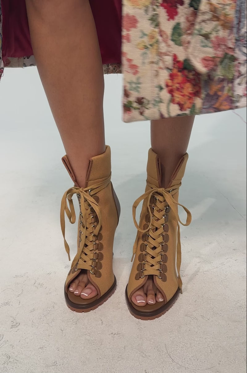 A video of a model wearing the AZALEA WANG PINGREE BROWN OPEN TOE WEDGE BOOTIE featuring a tan canvas upper, brown leather accents, lace-up front, peep toe, sculpted dark brown wedge heel, and a modern lug sole.