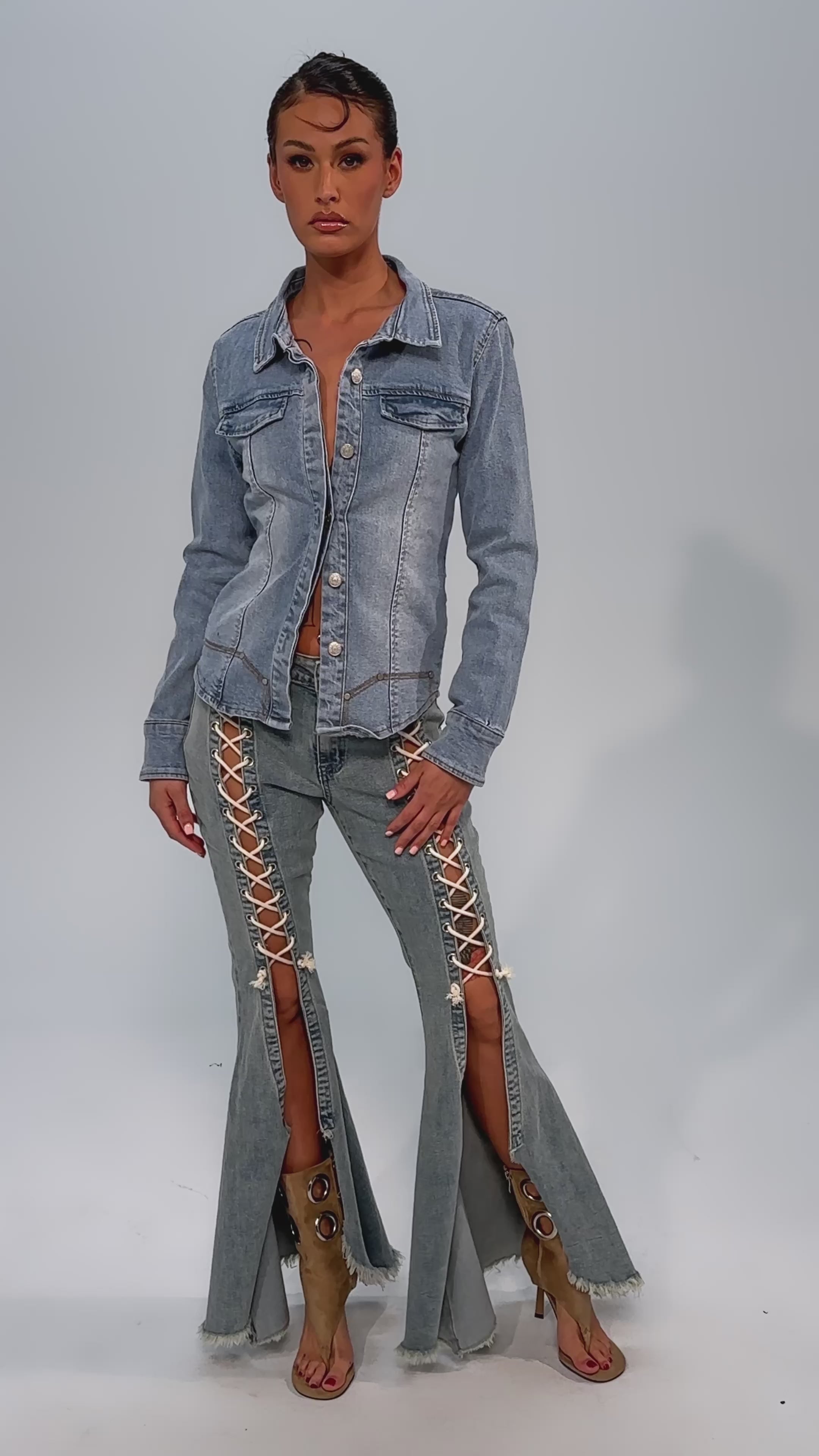 Video of woman modeling the CRAZY ENOUGH LACE UP FLARE DENIM, paired with a denim jacket, tan high-heeled boots, and a beige clutch. The pants have front slits and lace-up detailing. She stands against a plain light gray background.