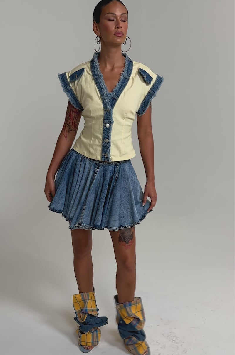 A video of a model in the DO YOU LOVE ME BUTTON DOWN, styled with a blue denim pleated mini skirt and plaid yellow-and-blue slouchy high-heeled boots.
